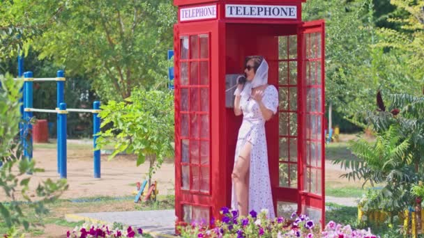 Güzel genç bir bayan, İngiliz tarzı kırmızı bir telefon kulübesinde mutlu bir şekilde konuşuyor. Beyaz elbiseli ve gözlüklü kız.. — Stok video