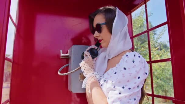 Die schöne junge Frau telefoniert fröhlich in einer roten Telefonzelle im englischen Stil. Mädchen in weißem Kleid und Brille. — Stockvideo