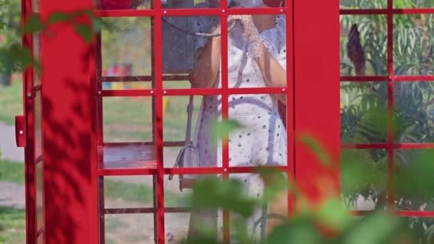 Bela jovem fala alegremente ao telefone em uma cabine de telefone vermelho estilo inglês. menina vestida com um vestido branco e óculos. — Vídeo de Stock