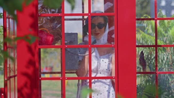 Güzel genç bir bayan, İngiliz tarzı kırmızı bir telefon kulübesinde mutlu bir şekilde konuşuyor. Beyaz elbiseli ve gözlüklü kız.. — Stok video