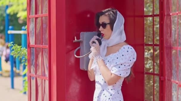 Bella giovane donna parla felicemente al telefono in stile inglese cabina telefonica rossa. ragazza vestita con un vestito bianco e occhiali. — Video Stock