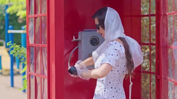美しい若い女性は喜んで英語のスタイルの赤い電話ブースで電話で話す。白いドレスを着た女の子と眼鏡をかけて. — ストック動画