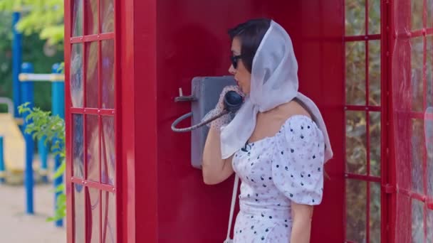 Die schöne junge Frau telefoniert fröhlich in einer roten Telefonzelle im englischen Stil. Mädchen in weißem Kleid und Brille. — Stockvideo