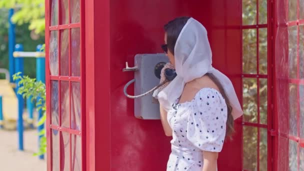 一位年轻漂亮的女士在一个英式的红色电话亭里愉快地通过电话交谈。穿着白色衣服和眼镜的女孩. — 图库视频影像