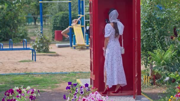 一位年轻漂亮的女士在一个英式的红色电话亭里愉快地通过电话交谈。穿着白色衣服和眼镜的女孩. — 图库视频影像