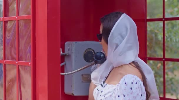 Bella giovane donna parla felicemente al telefono in stile inglese cabina telefonica rossa. ragazza vestita con un vestito bianco e occhiali. — Video Stock