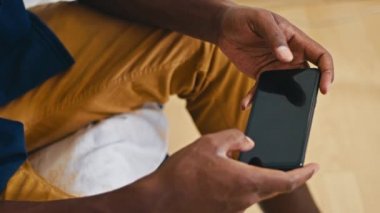 Mutlu Afrikalı adam yatakta oturuyor, kapüşonlusunu düzeltiyor, kameraya gülümsüyor, telefon mali istatistiklerine bakıyor..