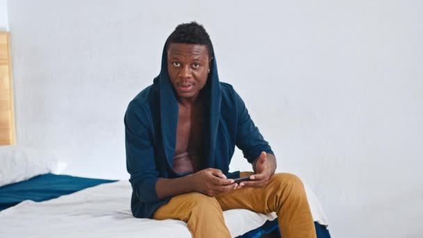 Glad afrikansk kille sitter på sängen, rätar ut sin luvtröja, ler mot kameran, tittar in i telefonens ekonomiska statistik. — Stockvideo
