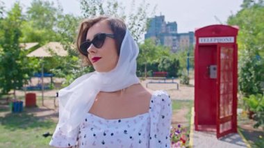 Mutlu genç bir kadın kırmızı bir telefon kulübesinin arka planında duruyor, gözlüklerini düzeltiyor. Etrafa bakıyorum. pinup.