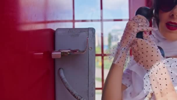 Όμορφη νεαρή γυναίκα μιλάει ευτυχισμένα στο τηλέφωνο σε ένα αγγλικό στυλ κόκκινο τηλεφωνικό θάλαμο. Ένα κορίτσι ντυμένο με λευκό φόρεμα και γυαλιά. pinup. — Αρχείο Βίντεο