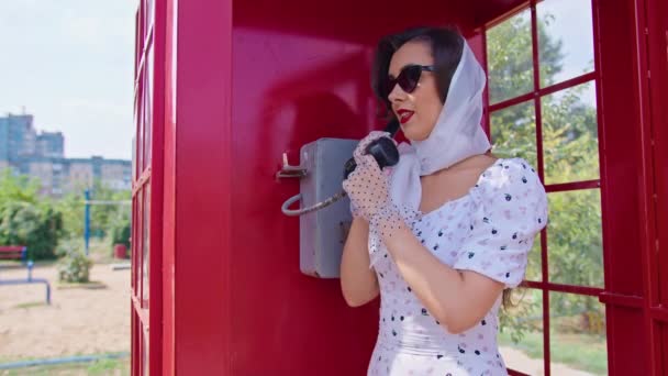Hermosa mujer joven felizmente habla por teléfono en una cabina telefónica roja de estilo Inglés. chica vestida con un vestido blanco y gafas. pinup. — Vídeo de stock