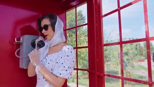 Die schöne junge Frau telefoniert fröhlich in einer roten Telefonzelle im englischen Stil. Mädchen in weißem Kleid und Brille. pinup. — Stockvideo