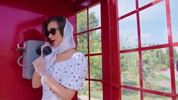 Bella giovane donna parla felicemente al telefono in stile inglese cabina telefonica rossa. ragazza vestita con un vestito bianco e occhiali. pinup. — Video Stock