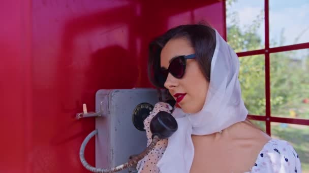 Bela jovem fala alegremente ao telefone em uma cabine de telefone vermelho estilo inglês. menina vestida com um vestido branco e óculos. pinup. — Vídeo de Stock