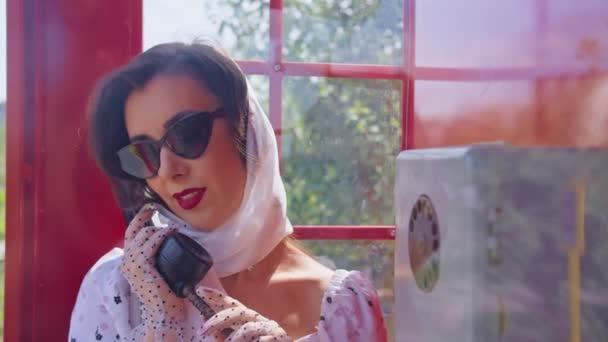 Bella giovane donna parla felicemente al telefono in stile inglese cabina telefonica rossa. ragazza vestita con un vestito bianco e occhiali. pinup. — Video Stock