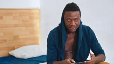 Mutlu Afrikalı adam yatakta oturuyor, kapüşonlusunu düzeltiyor, kameraya gülümsüyor, telefon mali istatistiklerine bakıyor..