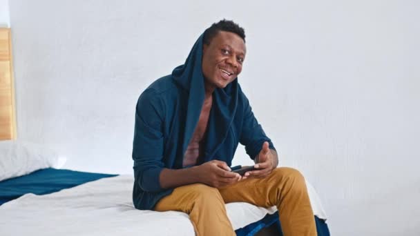 Feliz cara africano sentado na cama, endireita seu capuz, sorri para a câmera, olha para as estatísticas financeiras do telefone. — Vídeo de Stock