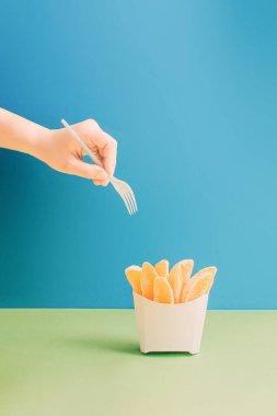 Yaratıcı sağlıklı yemek fikri, patates kızartması torbasında taze organik portakal dilimlerinin üzerinde çatal tutan kadınlar. Estetik diyet konsepti, parlak mavi ve yeşil pastel arka plan.