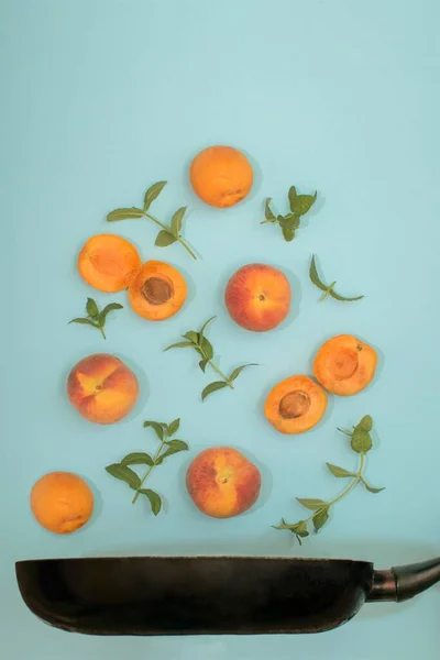 Minimalistic summer fruit idea with fresh organic apricot, peach and mint leaves above frying pan. Creative sweet and healthy food idea, aesthetic flat lay vertical arrangement, bright blue background.