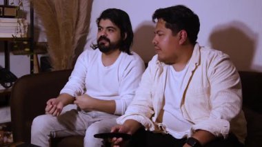 Uzun saçlı, sakallı, kanepeye oturmuş uzaktan kumandayla gülen İspanyol bir adamın videosu. Keyifli ev eğlencesi ve medya eğlencesi vurgulandı.