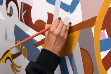 Fırçayı tutan ve kısmen bitmiş, soyut bir tuvale boya süren bir elin aşırı yakın çekimi. Sanatsal yaratma sürecinin titiz detayları, uygulamaları ve son aşamalarına odaklanır