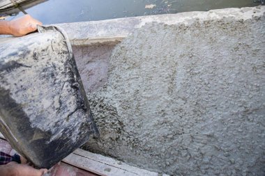 Fresh concrete mix being poured from bucket onto sand foundation near pond or river shore.