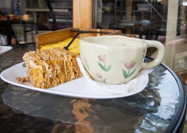 Cup of coffee and slice of honey cake on white plate served on glass table in cozy cafe. Concept of dessert, relaxation, and coffee break.