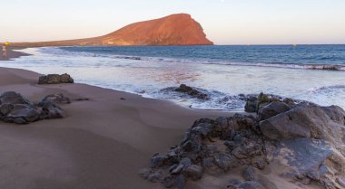Beautiful Montaa Roja beach in Tenerife with red volcanic mountain, ocean waves, and warm sunset light. Natural travel landscape and vacation destination.