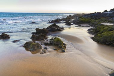 Güneş doğarken volkanik kayalar ve okyanus dalgalarıyla kumsal Tenerife, Kanarya Adaları, doğanın rahatlama ve güzelliğini simgeleyen doğal kıyı manzarası.