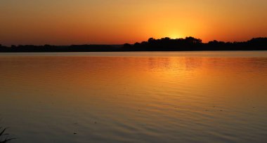 Turuncu gökyüzünde yansıması olan sakin nehrin üzerinde altın gün batımı huzurlu bir doğal manzara yaratır.
