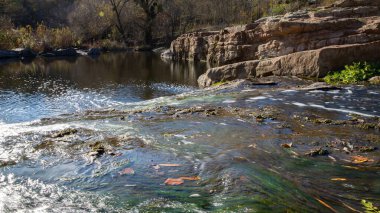 Nehir akıntısı güneş ışığında kayaların üzerinden akar. Su ve yeşillik ile doğal manzara.