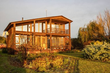 Günbatımının ılık ışığıyla aydınlatılmış geniş pencereli modern ahşap iki katlı bir ev.