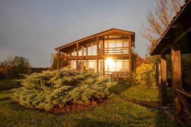 Günbatımının ılık ışığıyla aydınlatılmış geniş pencereli modern ahşap iki katlı bir ev.