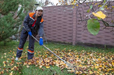 Evdeki bahçede çalışırken bir ağacın altında yaprakları tırmıklayan kadın.
