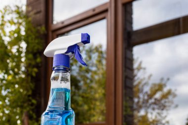 Gökyüzünü ve yeşil ağaçları yansıtan pencerenin yanında deterjan bulunan mavi temizlik spreyi şişesi.