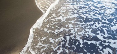 Islak siyah volkanik kumda okyanus köpük desenleri ve yumuşak kıyı şeridi eğrisi