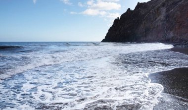 Okyanus dalgaları siyah volkanik kumsalları silip güneş ışığında kayalık yansıtıyor.