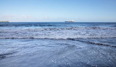 Okyanus dalgaları ufuktaki uzak kargo gemisiyle kayalık sahili yıkıyor.