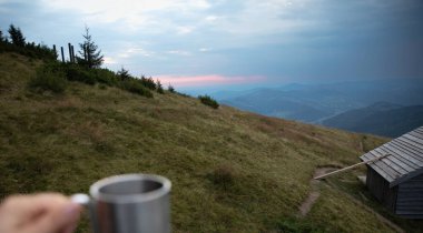 Elle tutulur metal kamp kupası sisli dağ manzarasına karşı ahşap kulübelerle