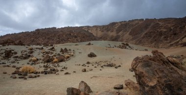 Dağılmış kayaları, kuru çalıları ve dramatik bulutlu gökyüzü olan çöl volkanik manzarası.