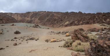 Dağılmış kayaları, kuru çalıları ve dramatik bulutlu gökyüzü olan çöl volkanik manzarası.
