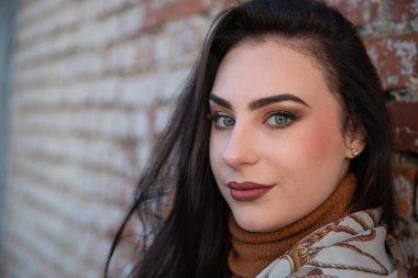 Güzel makyajlı genç bir kadın yumuşak akşam ışığında tuğla duvarın yanında poz veriyor.