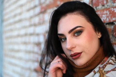 Güzel makyajlı genç bir kadın yumuşak akşam ışığında tuğla duvarın yanında poz veriyor.