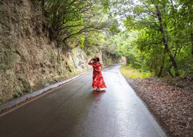 Çiçekli kırmızı elbiseli bir kadın Tenerife 'de seyahat ederken ıslak orman yolunda yürüyor.