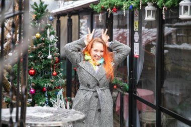 Neşeli kış gününde süslü Noel ağacının yanında komik bir hareketle poz veren neşeli genç kadın.