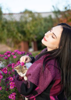 Genç bir kadın küçük bir kedi yavrusu tutuyor ve çiçeklere hafifçe dokunuyor.