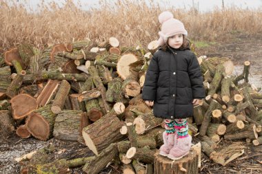 Küçük kız soğuk kış gününde odun yığınının yanında duruyor.
