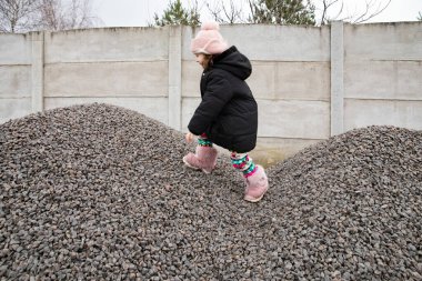 Soğuk kış gününde beton duvarın yanında çakıl tepesinde yürüyen küçük bir kız.