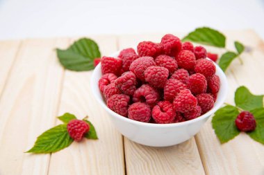 Taze olgun ahududular beyaz kasede, açık ahşap arka planda yapraklarla.