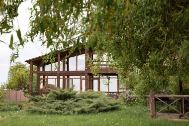 Bahçedeki büyük pencereli modern ahşap ev ağaç dallarının arasından görülüyor.